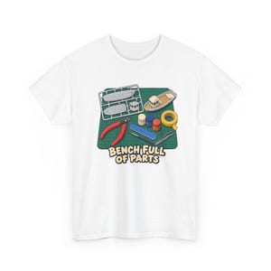 Könnte beinhalten: Ein weißes T-Shirt mit einer Grafik, die eine grüne Schneidematte mit Modellbootteilen, roten Seitenschneidern, Farbtöpfen, einem blauen Schleifstab, einem Pinsel, einer Pinzette und gelbem Klebeband zeigt, darunter der Text BENCH FULL OF PARTS.