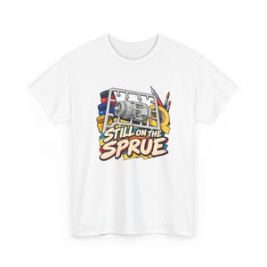 Könnte beinhalten: Ein weißes T-Shirt mit einer grafischen Illustration eines Modellbausatz-Spritzlings mit einem Strahltriebwerksteil, Farbtöpfen, Pinseln und Abklebeband. Der Text "STILL ON THE SPRUE" ist in fetten, stilisierten Buchstaben unter den Teilen gedruckt.