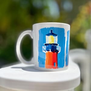 May include: White ceramic mug featuring a colorful illustration of a lighthouse. The lighthouse is painted with red, orange, blue, and yellow hues. The mug has a curved handle and is set against a blurred green background.