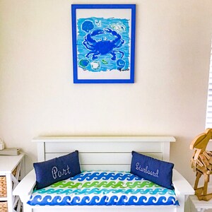 May include: White wooden bench with a blue and green wave pattern cushion. Two blue pillows with the words "Port" and "Starboard" are on the cushion. A framed blue crab painting hangs on the wall behind the bench.