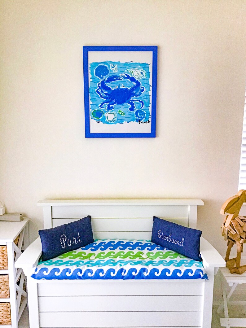 May include: White wooden bench with a blue and green wave pattern cushion. Two blue pillows with the words "Port" and "Starboard" are on the cushion. A framed blue crab painting hangs on the wall behind the bench.