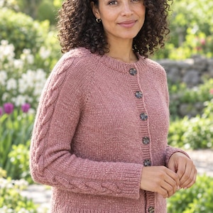 May include: A dusty rose knit cardigan featuring a crew neckline, long sleeves with vertical cable knit detailing, and a front button closure with five dark, mottled buttons. The garment is crafted from a textured, heathered yarn.