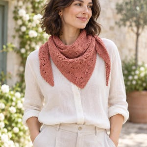 May include: A terracotta-colored triangular knit scarf featuring a delicate eyelet lace pattern along the edges. The scarf is worn over a long-sleeved white button-down shirt and light-colored trousers, showcasing the textured yarn construction.