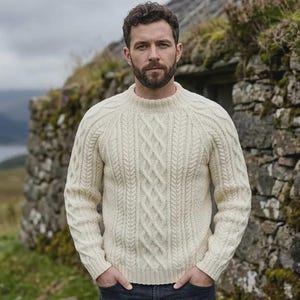 Könnte beinhalten: Ein cremefarbener Langarmpullover mit einem dicken Zopfstrickmuster, einem gerippten Rundhalsausschnitt und gerippten Bündchen. Das Kleidungsstück ist aus schwerem Strickmaterial gefertigt und verfügt über Raglanärmel für eine strukturierte Passform.
