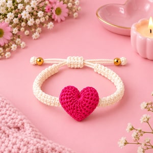 May include: A cream-colored macrame bracelet featuring a central bright fuchsia crocheted heart charm. The bracelet includes an adjustable sliding knot closure with two small, polished gold-tone metal beads at the ends of the cord strings.