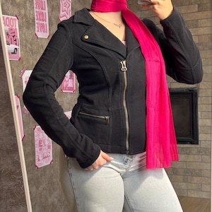 May include: A black long-sleeved jacket with a notched collar, a central gold-toned zipper, and a zippered pocket on the lower right side. A bright pink scarf is draped around the neck, hanging down the front. The jacket is worn with light blue jeans.