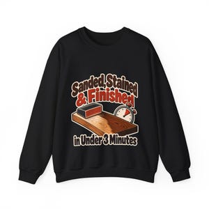 Peut inclure: Un sweat-shirt noir à col rond avec un motif représentant une planche en bois en cours de ponçage, un chronomètre et le texte "Sanded, Stained & Finished in Under 3 Minutes" imprimé en lettres marron, rouges et blanches sur la poitrine.