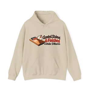 Peut inclure: Un sweat à capuche beige avec un motif représentant une planche en bois, un bloc de ponçage et un chronomètre. Le texte "Sanded, Stained & Finished in Under 3 Minutes" est imprimé en lettres marron et rouges au centre de la poitrine.