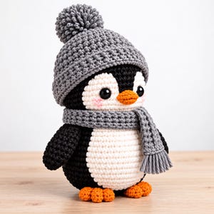 May include: A crocheted penguin toy featuring a black and white body, orange beak and feet, and a grey crocheted hat with a pom-pom on top. The penguin wears a matching grey crocheted scarf with fringe detailing and has small pink blush marks.
