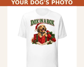 May include: A white t-shirt featuring the text DOX IN A BOX above a graphic of a light brown dachshund wearing a red Santa hat and a black collar, sitting inside a red and green Christmas gift box with a green ribbon and bow.