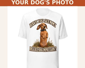 May include: A white short-sleeved t-shirt featuring a graphic of a brown dachshund wearing a black collar, standing upright in grass. The text above reads HAVE YOU SEEN MY and the text below reads PRAIRIE DOG IMPRESSION? in brown.