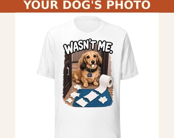May include: A white t-shirt featuring a graphic of a brown dachshund with a piece of toilet paper in its mouth, surrounded by scattered toilet paper on a blue mat, with the text WASN'T ME. above the dog and CUSTOMIZED WITH YOUR DOG'S PHOTO.