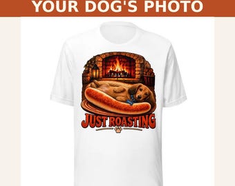 May include: A white t-shirt featuring a graphic of a brown dachshund lying inside a hot dog bun. The background shows a stone fireplace with a fire. Below the dog, the text reads JUST ROASTING in red letters with a small paw print icon.