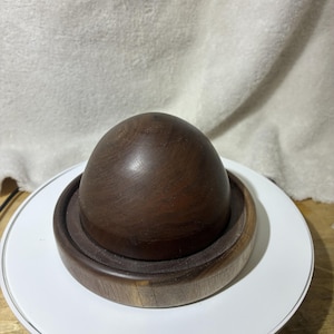 May include: A dark brown wooden dome-shaped object resting inside a matching circular wooden base. The wood features a visible natural grain pattern with a smooth, polished finish. The object is composed of two distinct parts, a rounded top piece and a shallow, flat-bottomed circular tray that supports the dome.