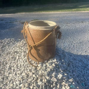 Op de afbeelding: Een cilindervormige bruine stenen pot met een getextureerd buitenoppervlak en een gladde, lichtgekleurde binnenrand. De pot is voorzien van twee zijdelingse oren met een dun, gevlochten beige touw als handvat om te dragen of op te hangen.