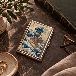 May include: A rectangular metal cigarette case featuring a printed illustration of a person in a patterned kimono surfing on a large blue wave, reminiscent of traditional Japanese woodblock art, with a polished silver-tone metal frame.