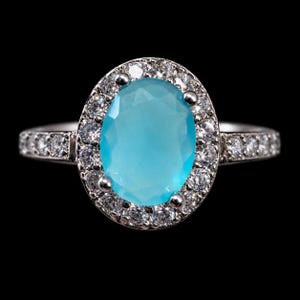 May include: A silver-toned ring featuring a large, oval-shaped, light blue center stone. The central stone is surrounded by a halo of small, clear, round-cut stones, with additional clear stones set along the band on both sides.
