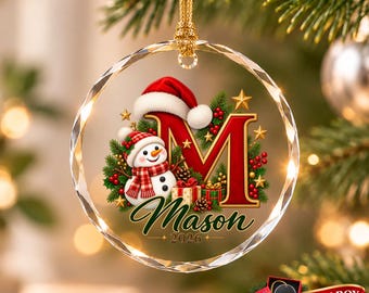 May include: A circular clear glass ornament featuring a large red letter M, a snowman wearing a red and white hat and scarf, green pine branches, red berries, and gift boxes. The text Mason and 2026 are printed below the letter M.