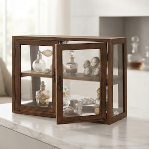 May include: A rectangular wooden display cabinet with glass panels and two hinged doors. The interior features two shelves holding various glass perfume bottles, small decorative items, and jewelry pieces. The frame is a medium-toned brown wood.