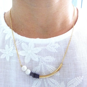 May include: A gold necklace with a curved bar pendant. The necklace has white and dark purple beads. It is worn over a white top with a floral pattern. The chain is delicate and gold-coloured.