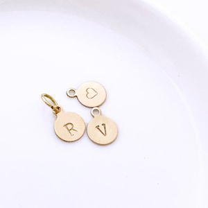 May include: Three small, gold-coloured charms on a white surface. Two round charms have the letters "R" and "V" engraved. The third charm is also round and features a heart. Each charm has a small loop for attaching to a chain or bracelet.