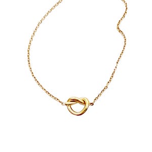 May include: A delicate gold-colored necklace featuring a knot pendant. The chain is thin and the knot is the focal point, creating a simple yet elegant design. The necklace is set against a white background.