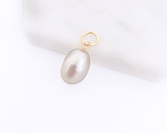 May include: A smooth, oval-shaped grey pearl pendant featuring a polished luster. The pearl is securely attached to a small, circular gold-toned metal jump ring at the top, designed for easy attachment to a necklace chain or bracelet link.