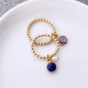 以下が含まれることがあります： 宝石チャームが付いた2つのゴールドビーズリング。1つのリングには紫のアメジストチャーム、もう1つには青いラピスラズリチャームと小さな白い真珠が付いています。リングは白い表面に表示されています。