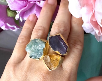 May include: A hand wearing three gold-plated rings, each featuring a different rough-cut gemstone: a light blue, a deep purple, and a golden yellow. The rings are set against a backdrop of pink and purple flowers.
