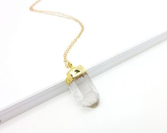 May include: A clear quartz crystal pendant necklace with a gold-colored cap and chain. The crystal is a natural, elongated shape. The chain is delicate and hangs from the top of the crystal.