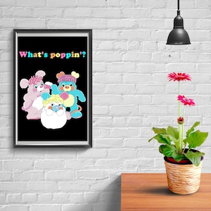 May include: A framed print featuring cartoon characters in pink, blue, and yellow on a black background. The text "What's poppin'?" is displayed in colorful letters. A potted plant with red flowers sits on a wooden surface.