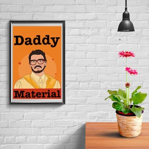 Puede incluir: Un póster enmarcado con las palabras "Daddy Material" arriba y abajo de una ilustración de dibujos animados de un hombre con gafas y un chal amarillo. Una planta en maceta con flores rosas está sobre una mesa de madera.