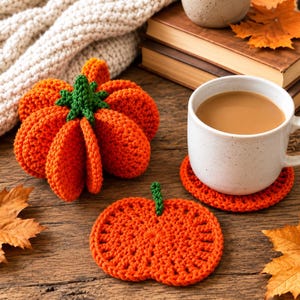 May include: A three-dimensional crocheted orange pumpkin with a green stem, and a flat, circular orange crocheted coaster with a small green stem, positioned next to a white ceramic mug filled with a light brown beverage.