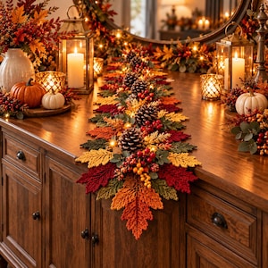 May include: A long, decorative table runner made of crocheted autumn leaves in shades of red, orange, yellow, and green. The runner is adorned with pinecones, red berries, and small fairy lights, creating a textured, seasonal display.