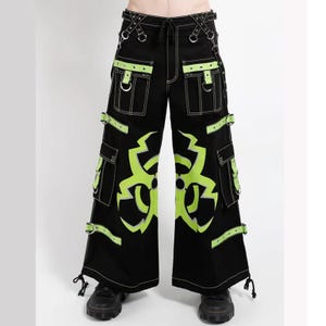 May include: Black wide-leg pants featuring neon green biohazard symbols on the front, multiple cargo pockets with straps, silver metal D-rings, and white contrast stitching. The pants include adjustable side straps and drawstring leg openings.