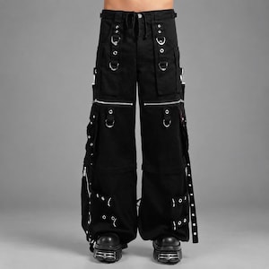 May include: Black cargo pants featuring multiple silver-toned metal D-rings, eyelets, and zippers. The trousers include large side pockets, adjustable straps with buckles, and a drawstring waist, providing a utilitarian design with wide legs.