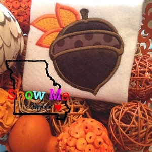 May include: A brown and white acorn with orange leaves, embroidered on a white fabric background. The fabric is surrounded by brown wicker balls and orange decorative balls.