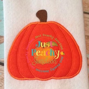 May include: Orange felt pumpkin with a stitched design. The pumpkin has a brown stem and a circle in the center with the text "Just Peachy Junction" in colorful letters.