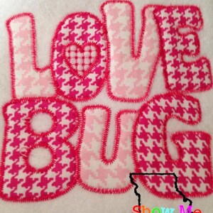 May include: Pink and white fabric letters spelling "LOVE BUG" with a small pink heart in the center of the "O". The letters are sewn with a red thread and have a houndstooth pattern.