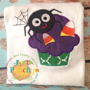 May include: A black spider with a web sits on top of a purple cupcake with green frosting and candy corn decorations. The cupcake is embroidered on a white t-shirt. The text "Just Peachy Junction" is embroidered on the bottom of the shirt.