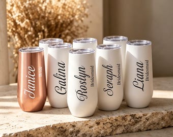 May include: A collection of six stainless steel stemless champagne flutes with clear lids. One flute is metallic rose gold with the name Janice and Bride printed in white. Five white flutes feature black names and the title Bridesmaid.