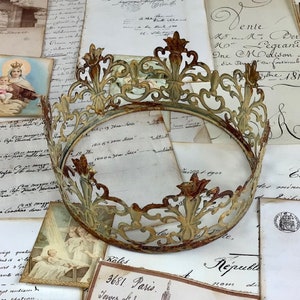 May include: An antique, openwork metal crown with a distressed, aged finish. The crown is circular with ornate floral and fleur-de-lis designs. It rests on a backdrop of vintage documents and religious imagery.