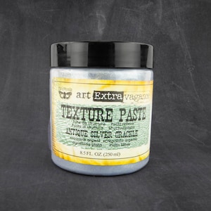 May include: A silver jar of texture paste labeled "art Extra vagance Texture Paste Antique Silver Crackle" with the text "Pôte de structure Pasta relieve Pasta di struttura Structuurpasta craquelé argent screpolata argento agrietado plata Platin Silber 8.5 FL. OZ. (250 ml)"