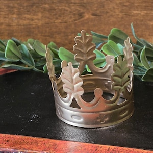 May include: A gold-colored metal crown with a decorative leaf design. The crown is made of thin metal and has a simple, elegant design.