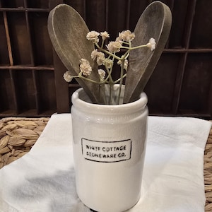 May include: A white stoneware jar with the text "WHITE COTTAGE STONEWARE CO." in a black rectangle. Two wooden spoons and small beige flowers are inside the jar. The jar sits on a white cloth.