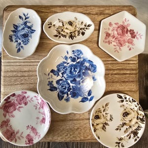 May include: A collection of decorative ceramic dishes with floral designs. The dishes are in various shapes, including oval, round, and hexagonal. The floral patterns feature blue, pink, and brown roses and foliage against a white background.