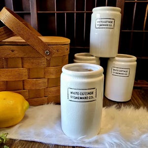 May include: A collection of white ceramic jars with the text "WHITE COTTAGE STONEWARE CO." in black. The jars are cylindrical with a slightly textured surface. A wooden picnic basket and a lemon are also visible.
