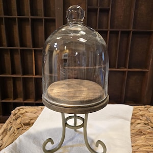 May include: A clear glass cloche dome with a round knob handle sits atop a circular dark wood base. The base is supported by a light-colored metal stand featuring three curved legs that terminate in scroll-shaped feet at the bottom.