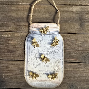 May include: A decorative mason jar-shaped wall hanging with a light blue and white painted finish. The jar is adorned with six gold-colored bee embellishments and a jute rope handle. The background is a rustic wooden surface.
