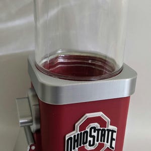 May include: A red and silver candy dispenser featuring a clear glass top container and the Ohio State logo on the front. The device has a silver base and a silver dispensing handle on the side, designed for holding and releasing small items.